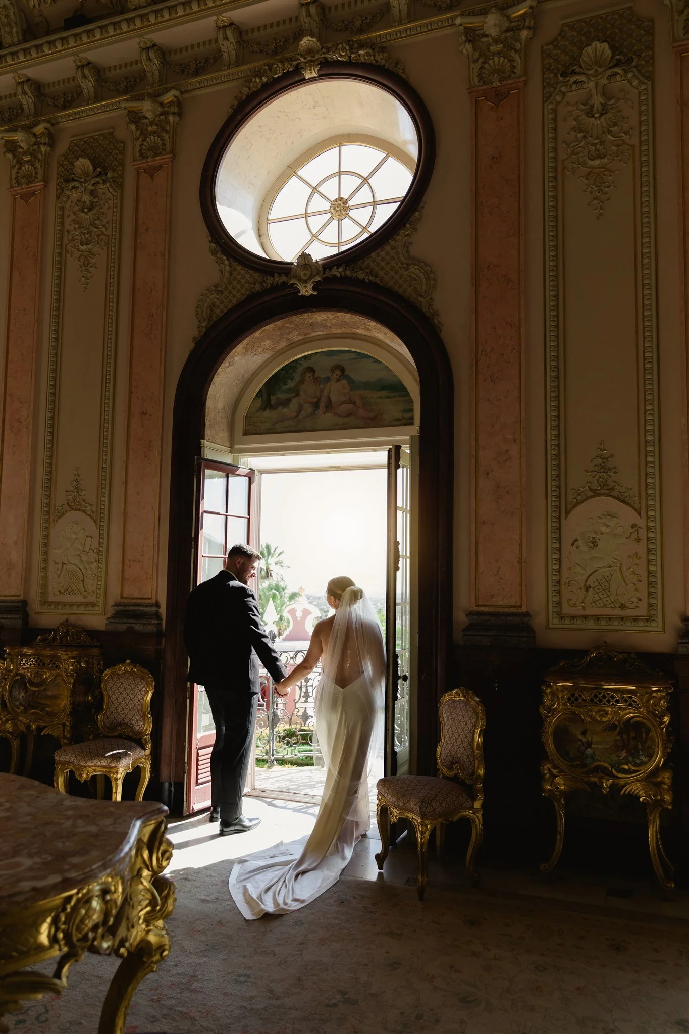 A bride and groom holding hands inside a lavish, ornate room with gold-trimmed furniture and decorative wall art, standing near an open window with a view of palm trees outside.