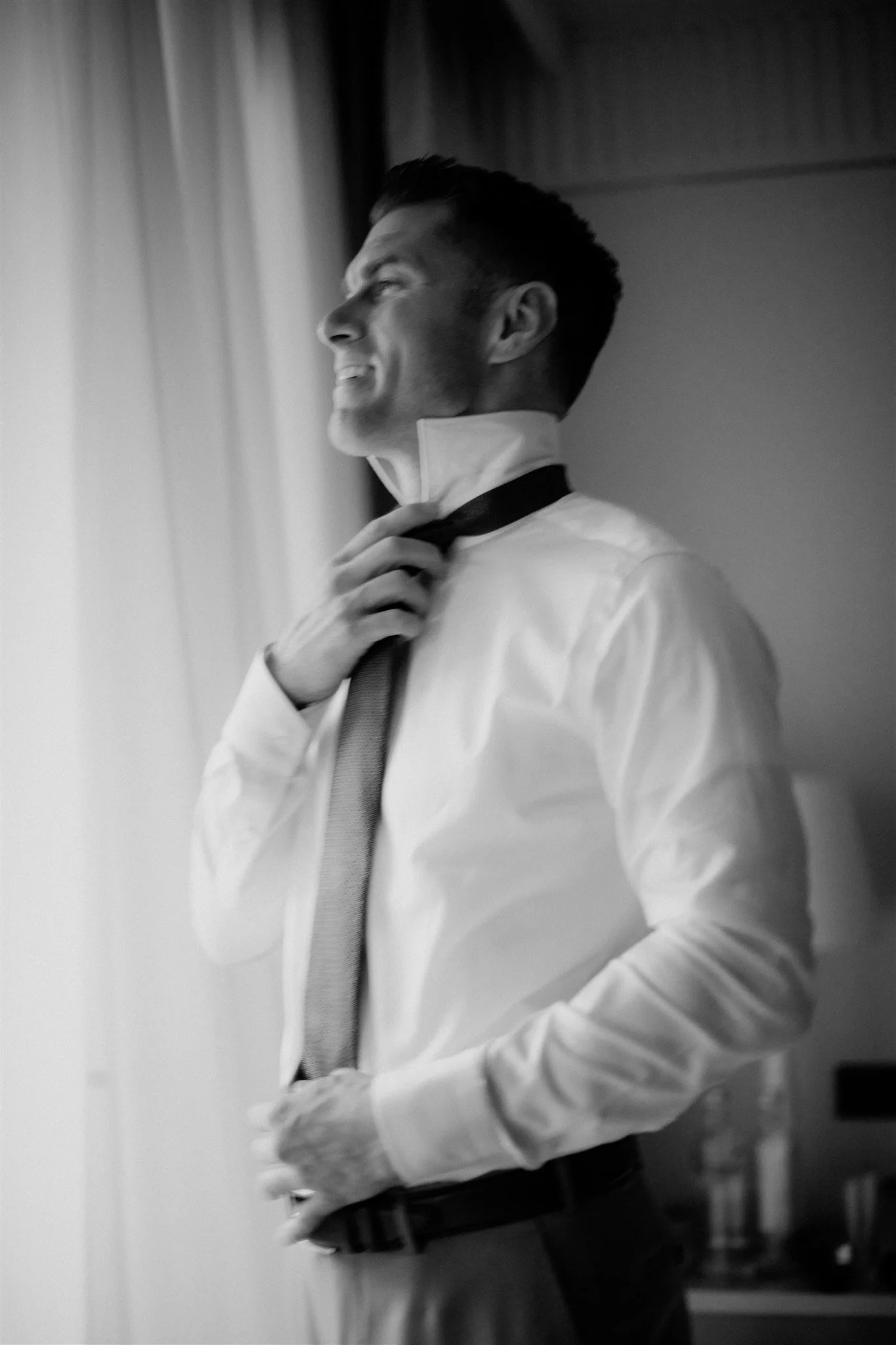 Black and white photo of a man in formal attire adjusting his necktie, standing indoors near a window with a curtain.