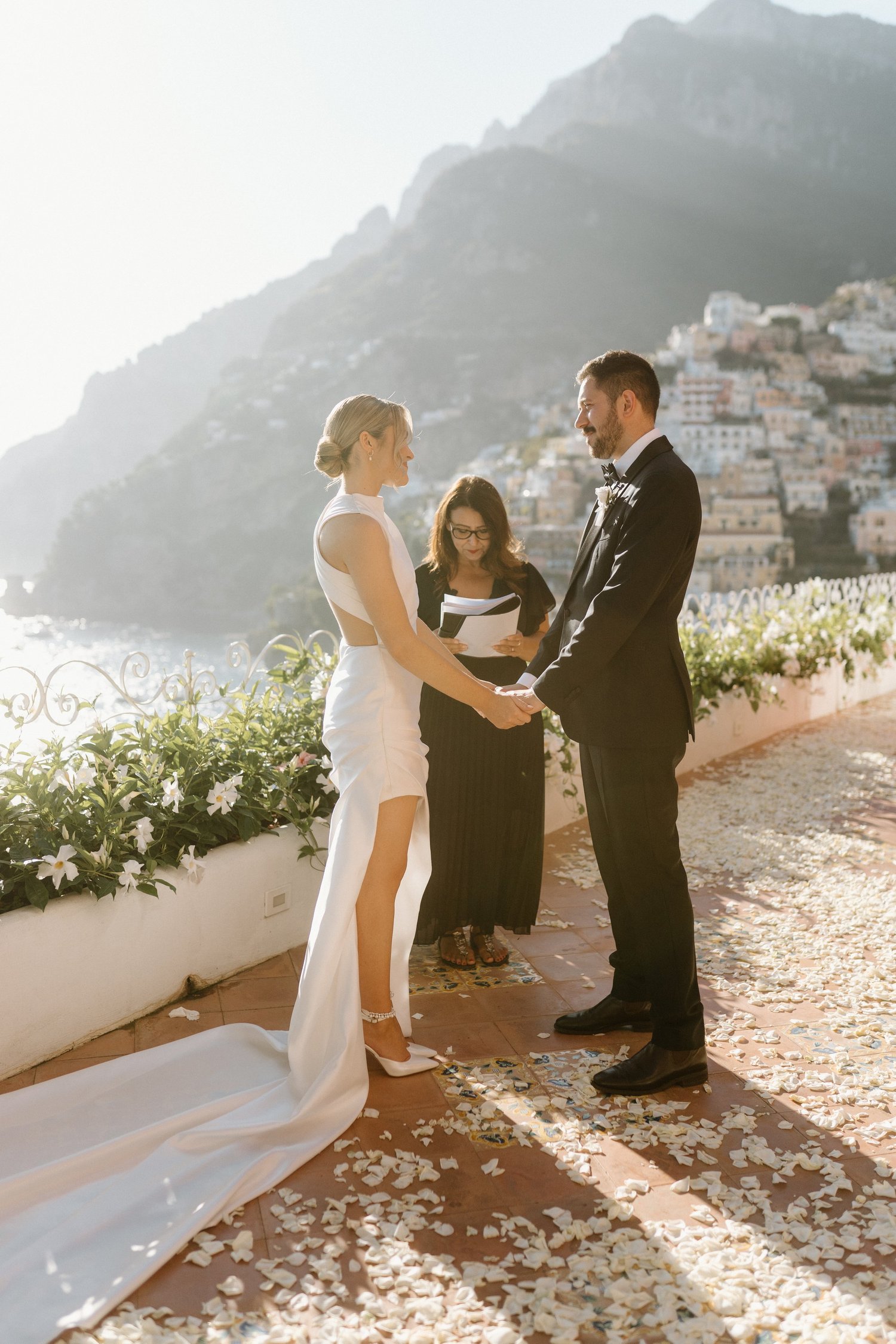 A bride and groom holding hands during wedding ceremony on an outdoor terrace overlooking seaside village and mountains, with an officiant officiating.