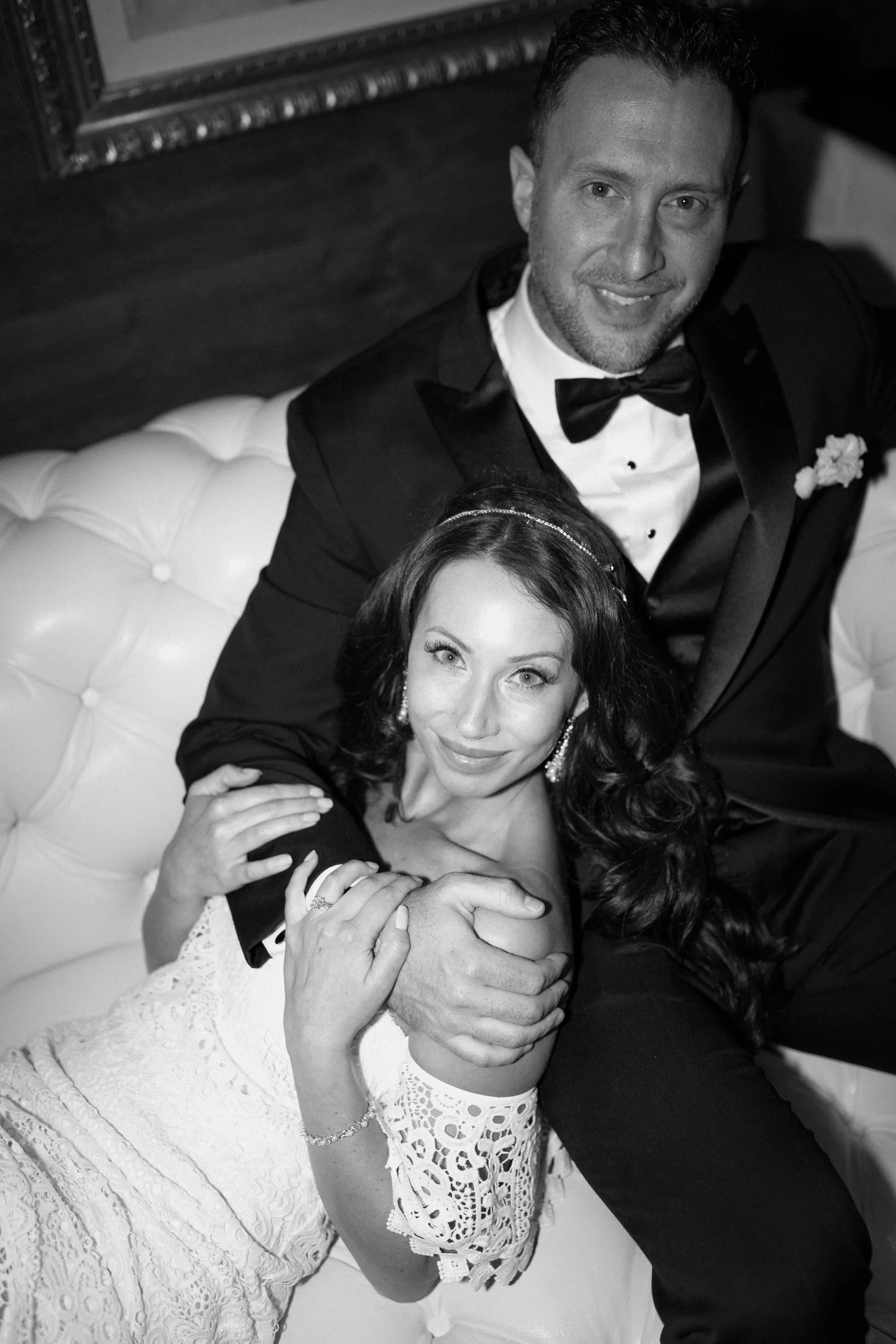 Black and white photo of a smiling man in formal tuxedo with bow tie and boutonniere, and a woman with long dark hair in a lace dress, embracing and looking up at the camera.