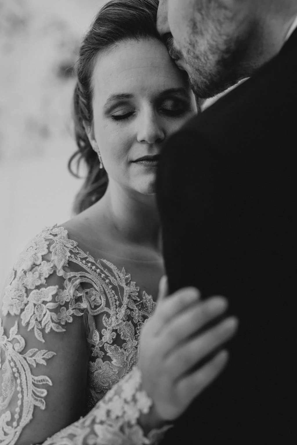 Black and white photo of a bride and groom sharing a tender moment, with the groom kissing the bride's forehead, her eyes closed and showing lace wedding dress details.