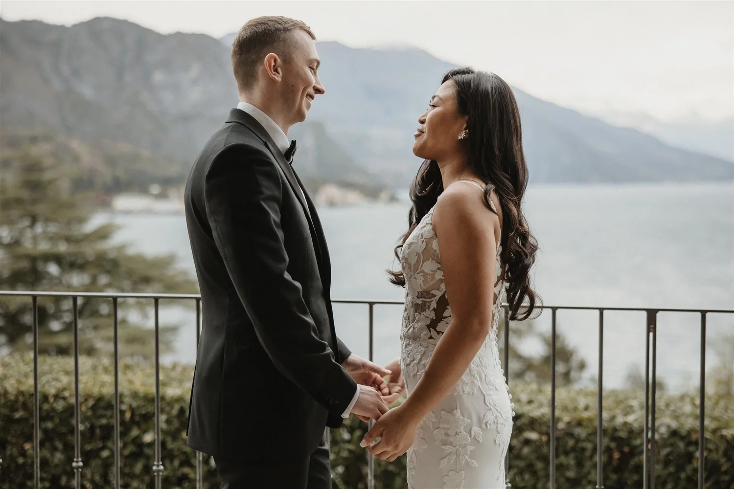 A bride and groom holding hands while facing each other outdoors with mountains and a lake in the background.