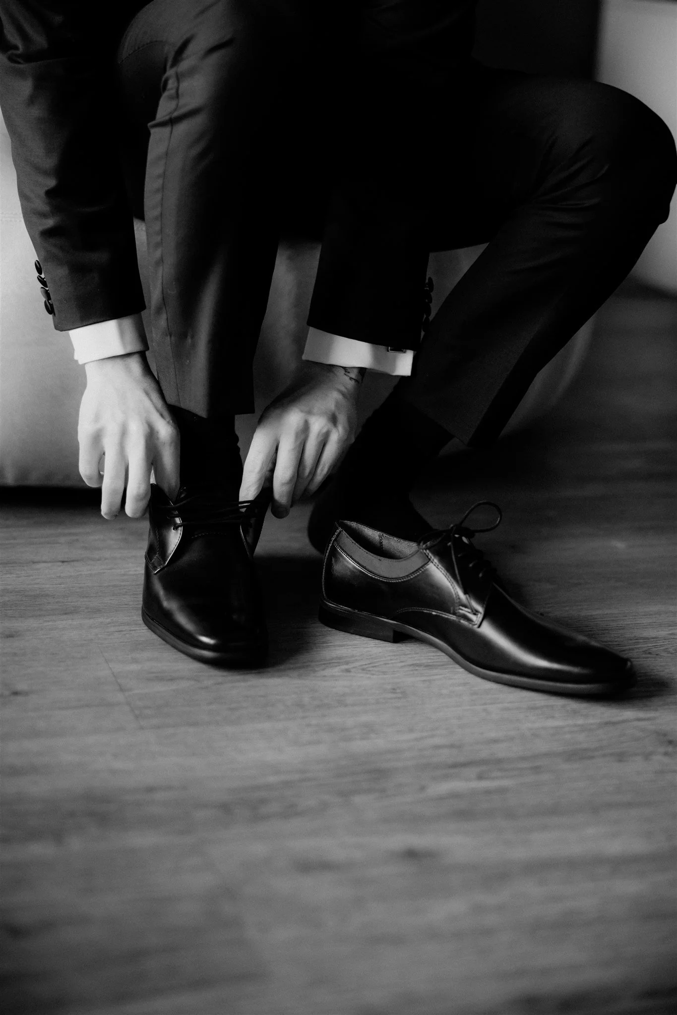 A person dressed in a suit is kneeling on the floor, tying their black dress shoes. The person is wearing dark pants and a suit jacket, with shirt cuffs visible. The floor is wooden, and the background is plain.