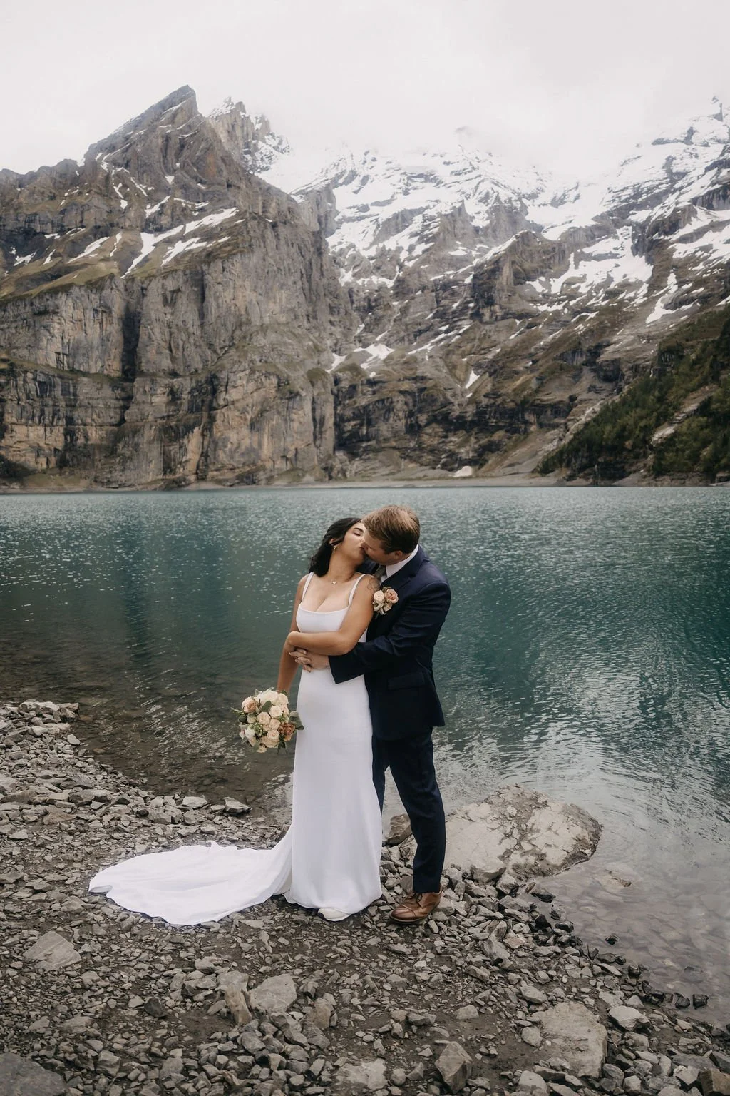 A newlywed couple embracing and kissing by a mountain lake, with snow-capped peaks and rugged cliffs in the background.