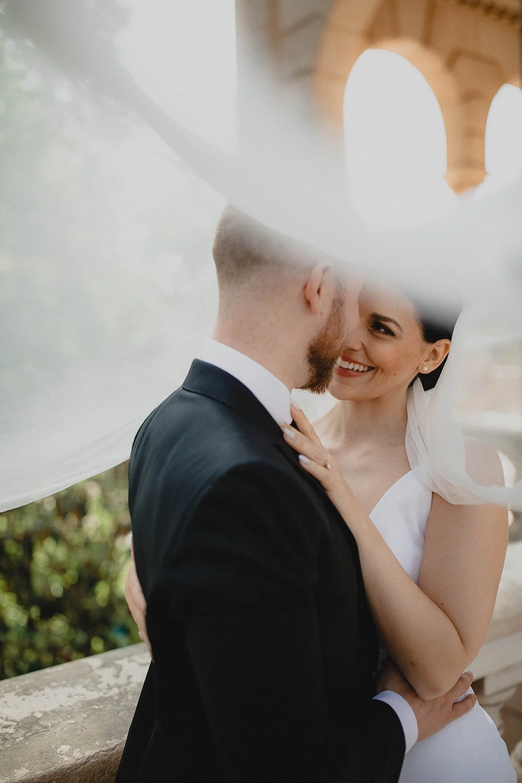 A bride and groom share a kiss under a veil on their wedding day, smiling at each other.
