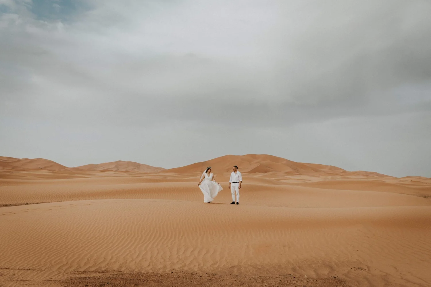 A couple walking together in a desert with sand dunes under a cloudy sky.