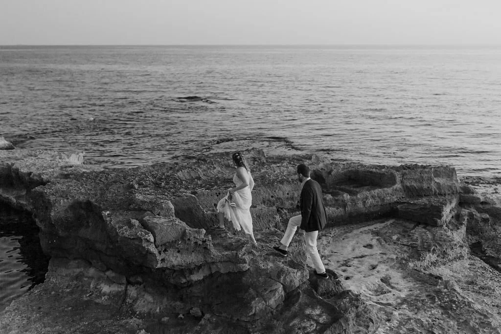 A black and white photo of a couple by the sea, with the woman walking on rocks and the man following behind, near the shoreline.