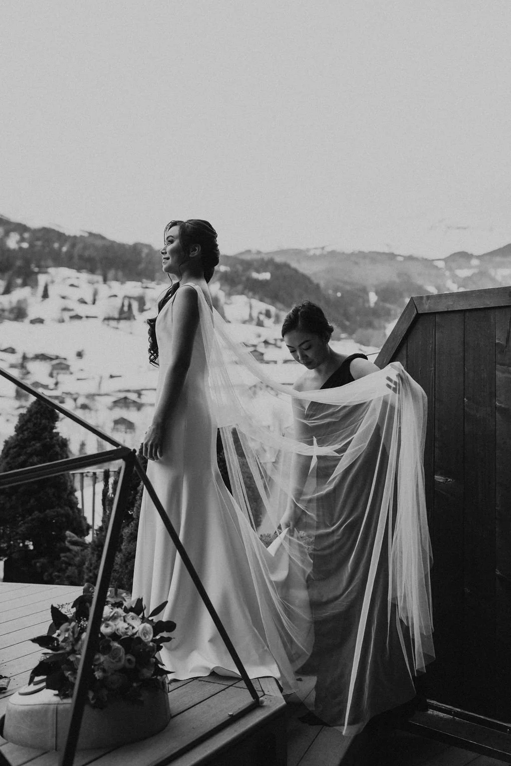 A bride in a wedding dress is being helped with her veil by a woman, on an outdoor balcony with a mountain landscape in the background.