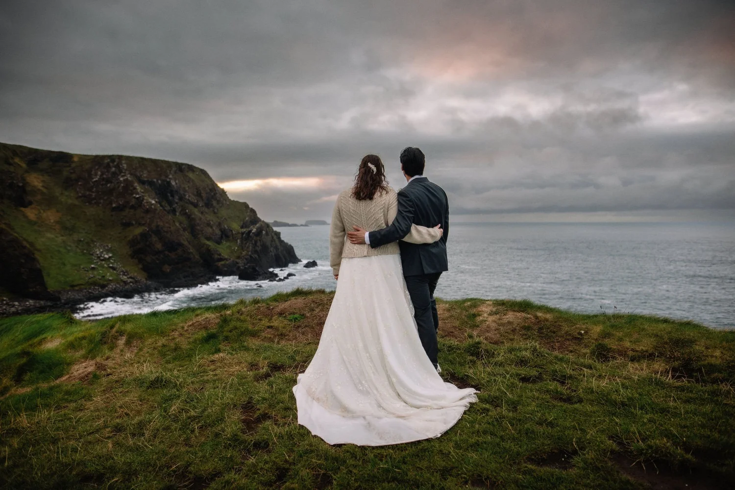 A couple dressed in wedding attire, standing on a grassy cliffside overlooking the ocean with rocky cliffs, under a cloudy sky at sunset.