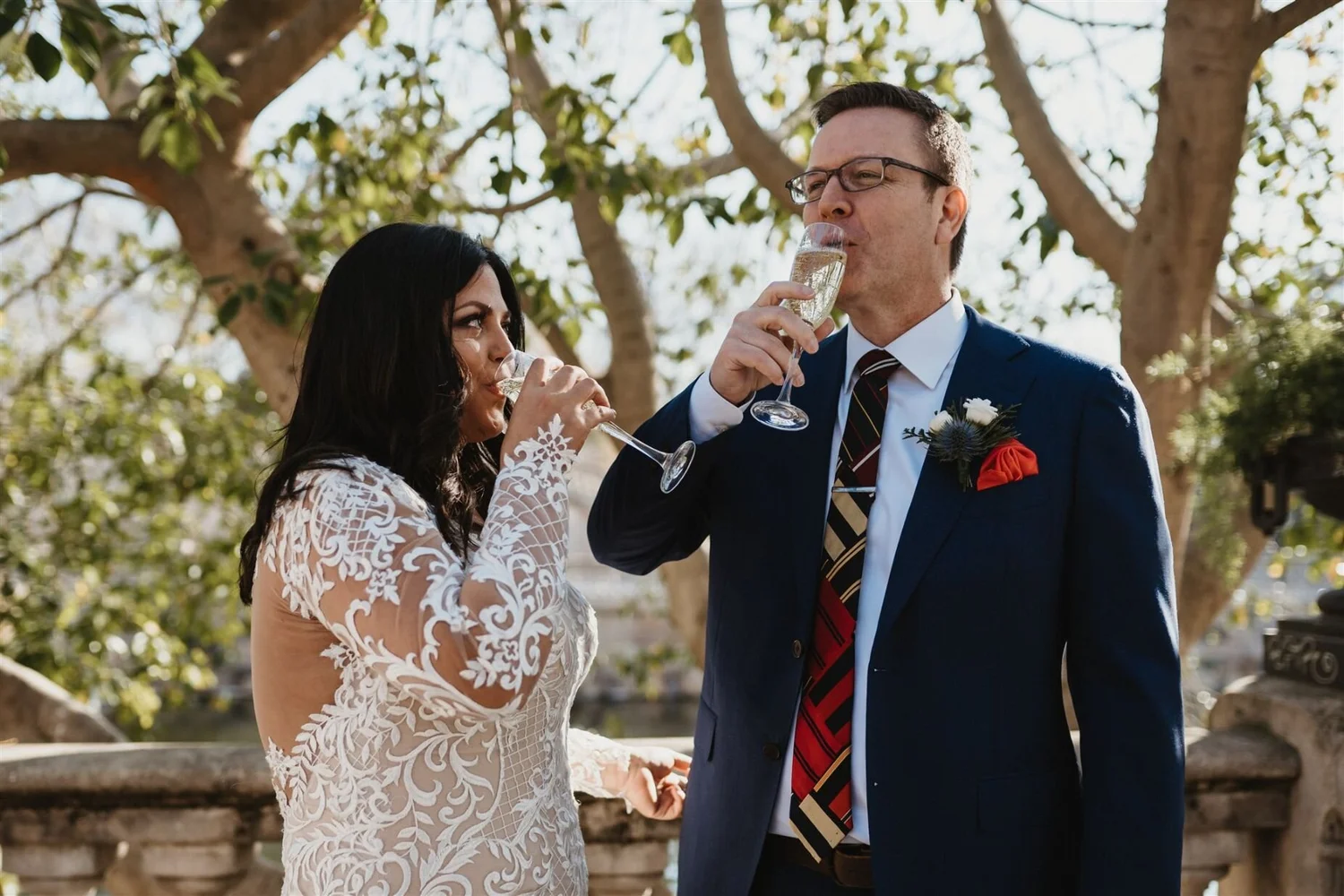 Bride and groom wearing formal attire, drinking champagne outdoors during their wedding ceremony with trees in the background.