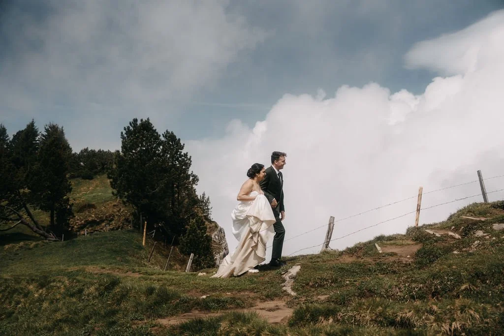 A bride and groom walking on a dirt path in a grassy, mountainous area during cloudy weather
