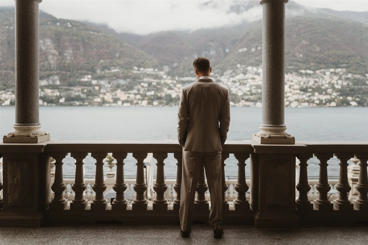 A man in a beige suit standing on a balcony, looking out over a lake with mountains in the background, framed by two stone columns.