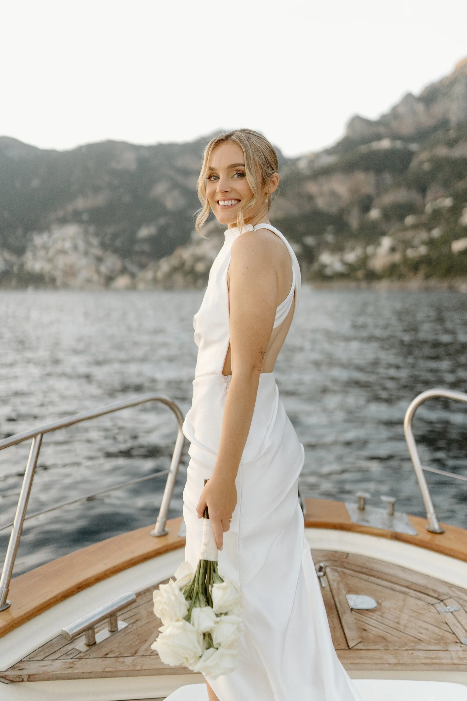 A woman in a white dress holding a bouquet of white roses on a boat with a mountainous landscape in the background.