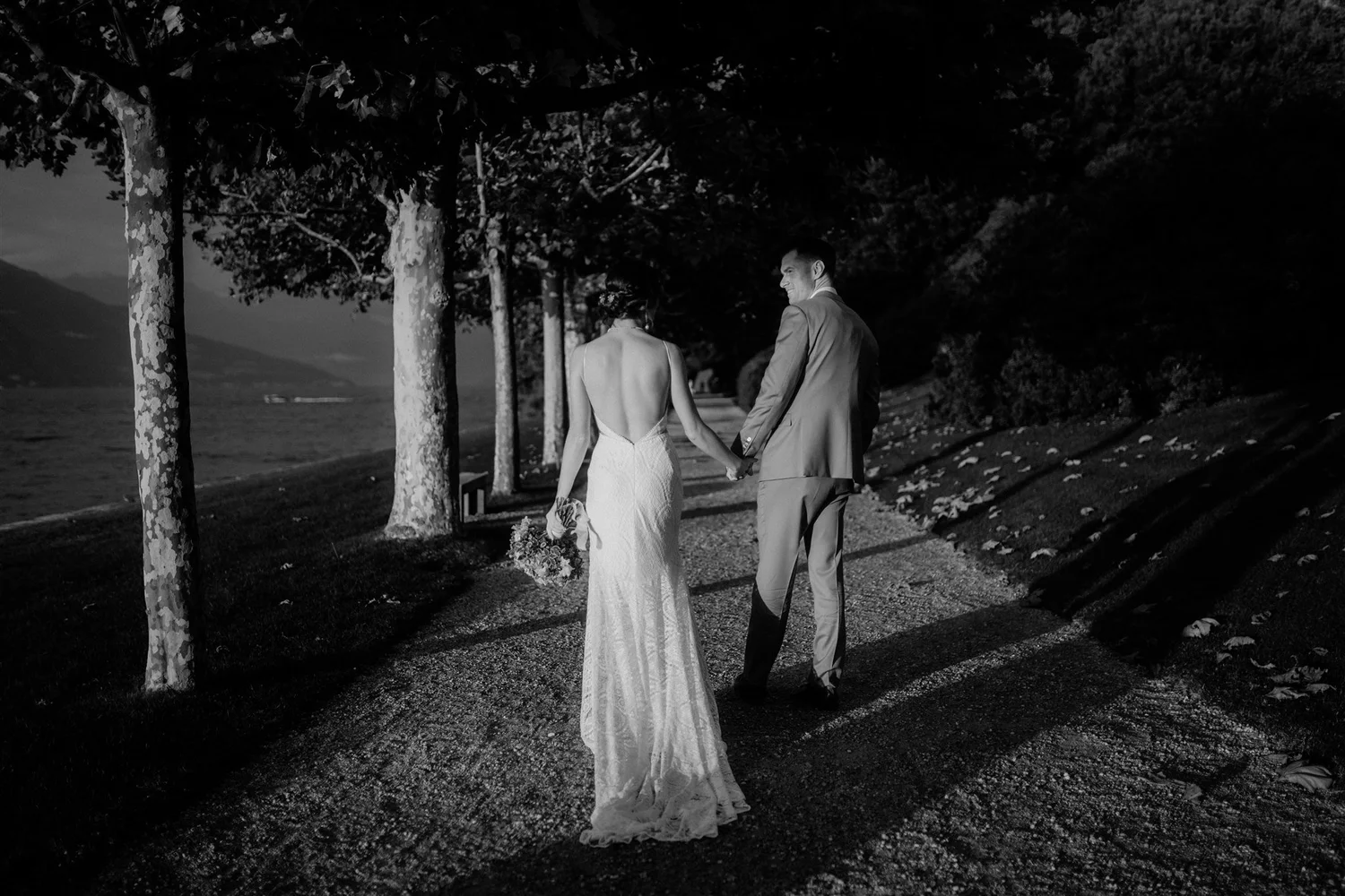 A black and white photo of a bride and groom holding hands and walking along a tree-lined path by a body of water during sunset or sunrise, with the groom smiling and the bride holding a bouquet.