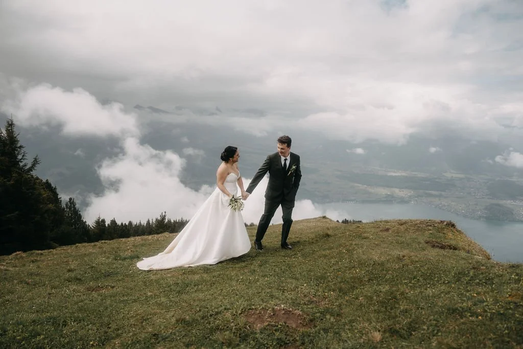 A bride and groom holding hands on a grassy hilltop with clouds, mountains, and a lake in the background.