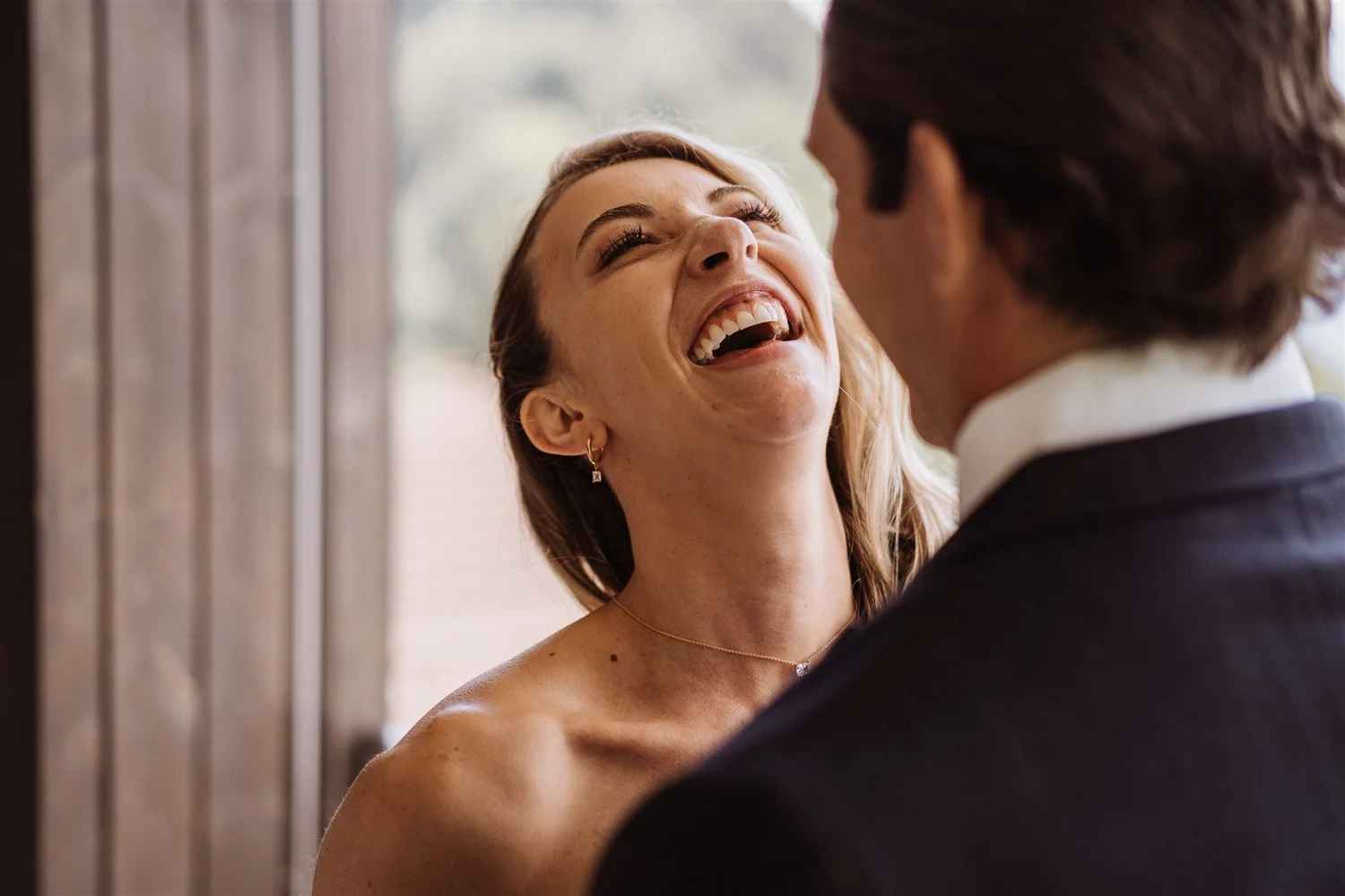 A woman with blonde hair smiling and looking happy, leaning toward a man with dark hair. She is wearing earrings and a necklace, and there is a window with blinds in the background.