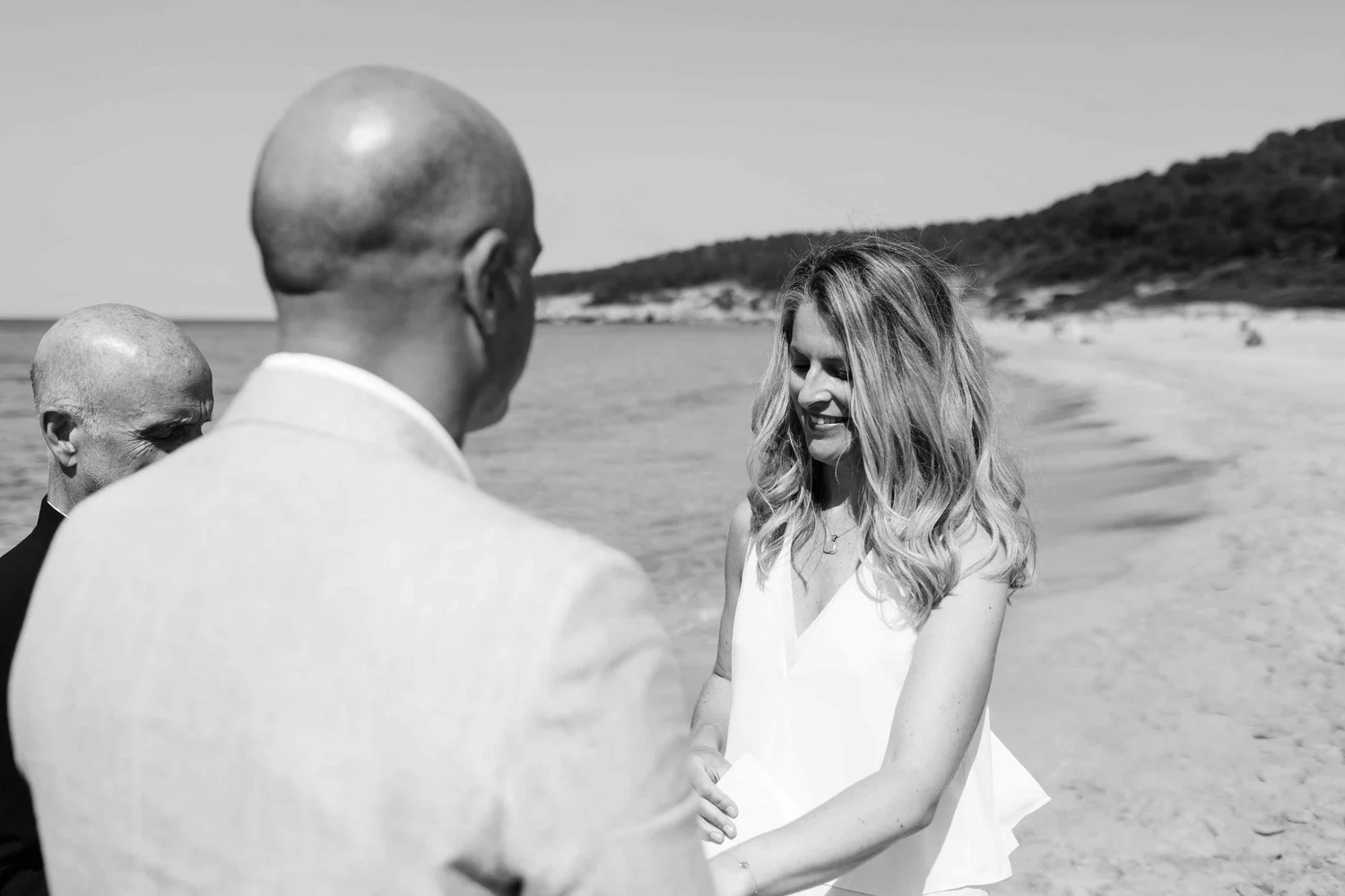 A black and white photo of a woman in a white dress smiling and holding hands with a man at a beach, with another man partially visible and an ocean and hillside in the background.