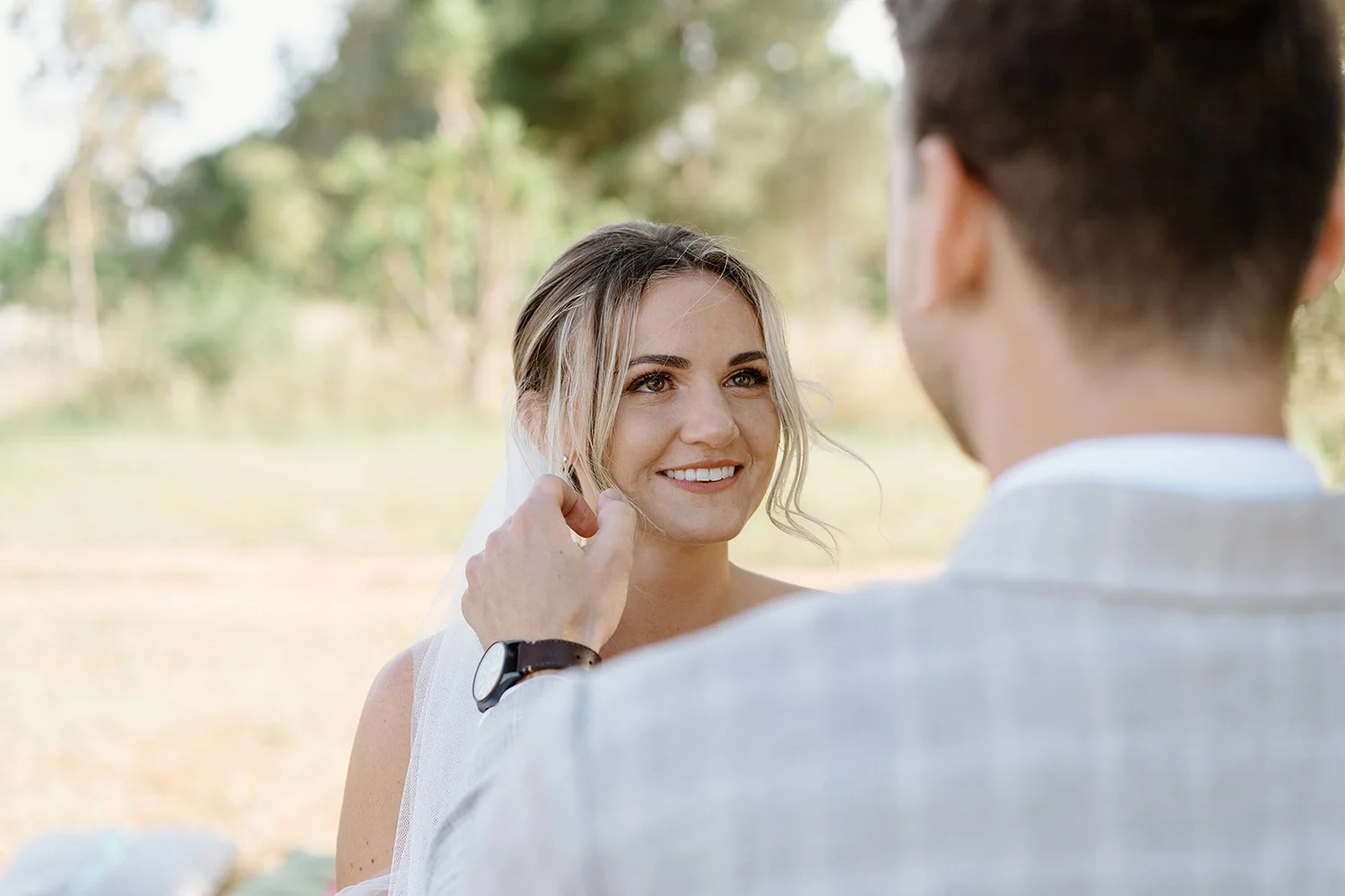 A bride with blonde hair and a smile looking at a man, who is holding her face gently and wearing a suit and watch, outdoors with a blurred background of trees.