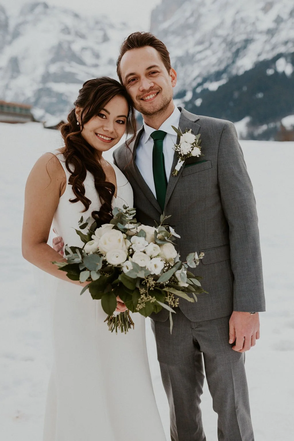 A newlywed couple standing outdoors in the snow with snowy mountains in the background. The bride is holding a bouquet of white flowers and greenery, wearing a white wedding dress with long dark hair. The groom is wearing a gray suit with a green tie