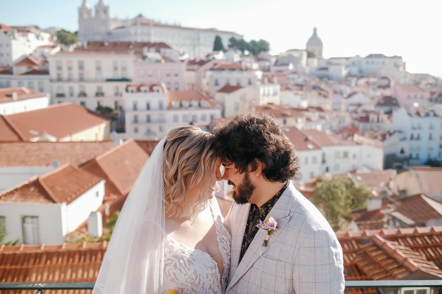 A bride and groom face each other with foreheads touching on a rooftop with a cityscape of red-tiled roofs and historic buildings in the background.