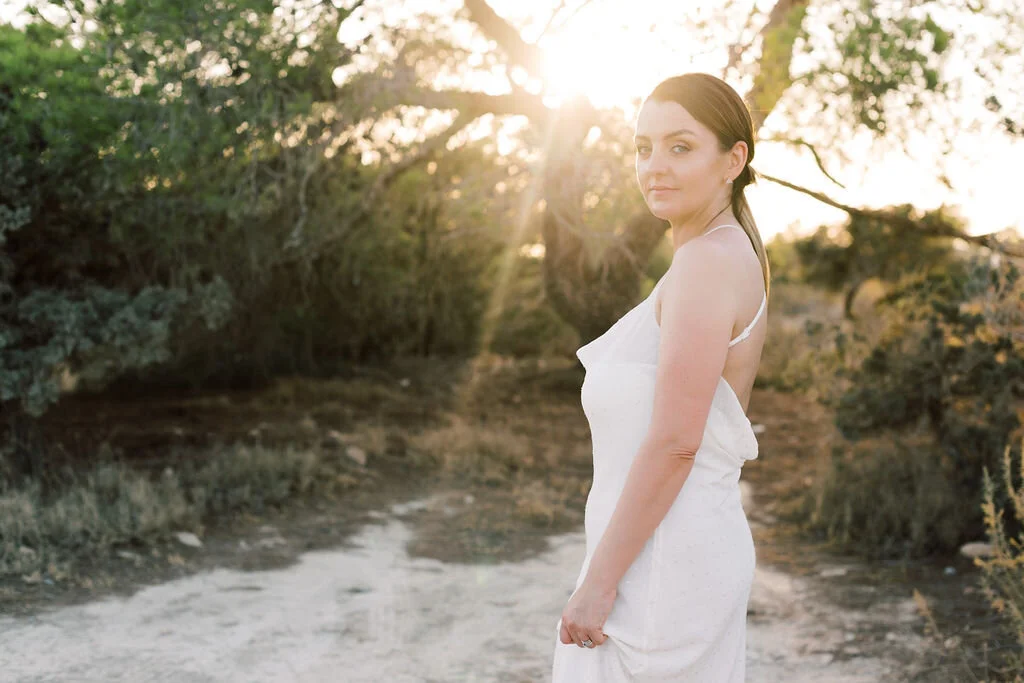 A woman standing outdoors in a white dress with sunlight shining through trees in the background.