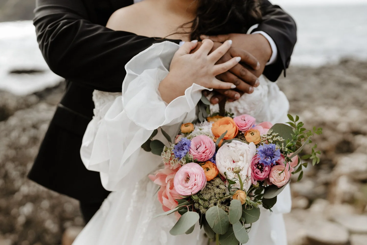 Couple embracing on a beach, with the bride holding a bouquet of pink, orange, purple, and white flowers.