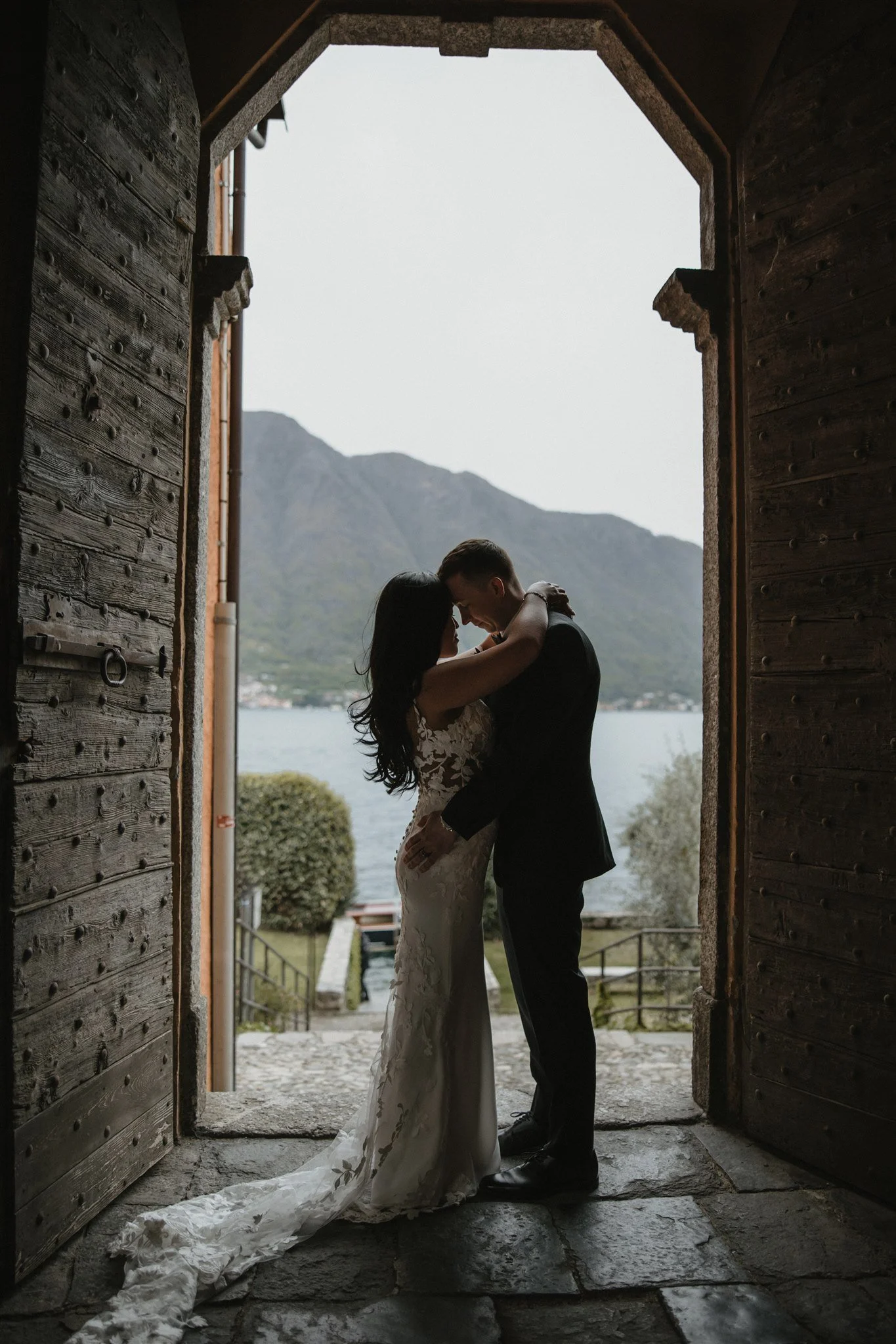 A newlywed couple sharing a romantic moment, embracing in a doorway with mountains and water in the background.