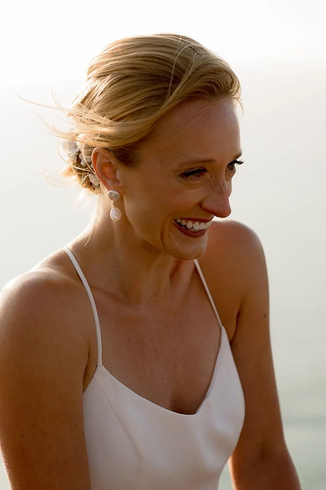 A woman with blonde hair in a white dress smiling at the beach during sunset.