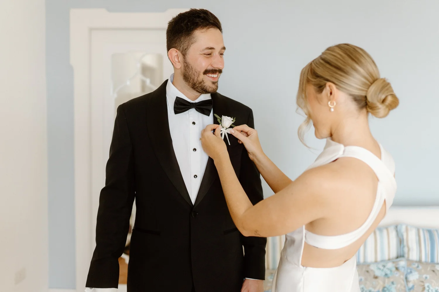 A man dressed in a black tuxedo with a bow tie and a woman in a white wedding dress sharing a moment during a wedding ceremony or preparation.