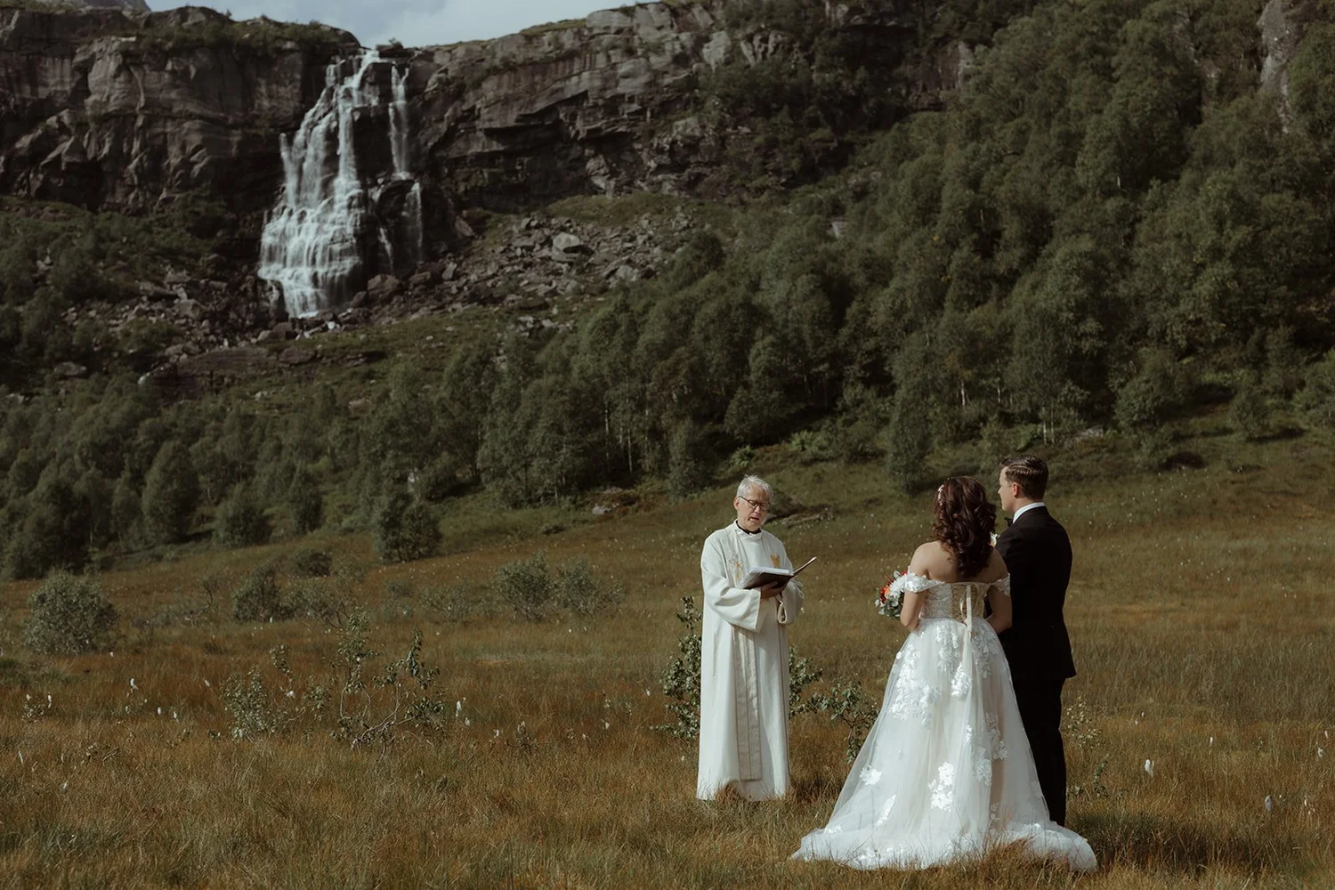 A wedding ceremony taking place outdoors in a scenic mountain landscape with a waterfall in the background. The bride and groom stand together, facing an officiant who is reading from a book. The bride is wearing a white wedding dress with ruffled of