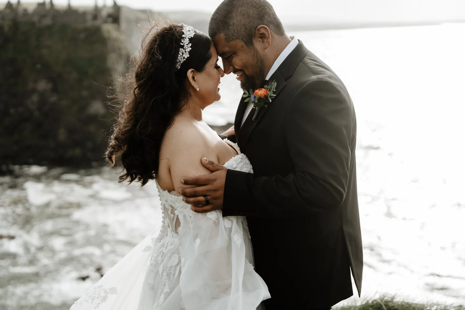 A bride and groom share a kiss outdoors near a body of water, with their foreheads touching and eyes closed, dressed in wedding attire.