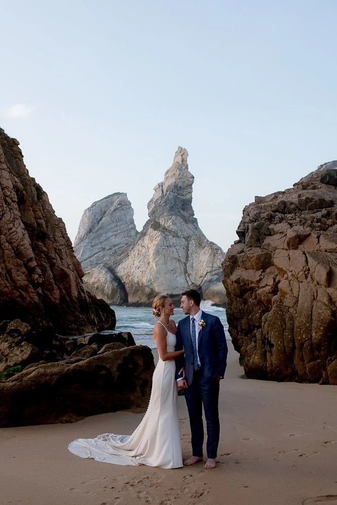 A bride and groom holding hands on a sandy beach, with large rocks and ocean behind them, and a tall rock formation in the distance.