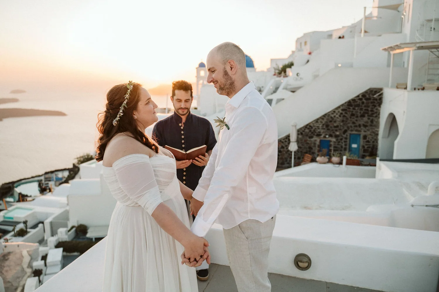 A couple getting married outdoors at sunset in Santorini, Greece, holding hands and gazing at each other with an officiant behind them, with white buildings and the sea in the background.