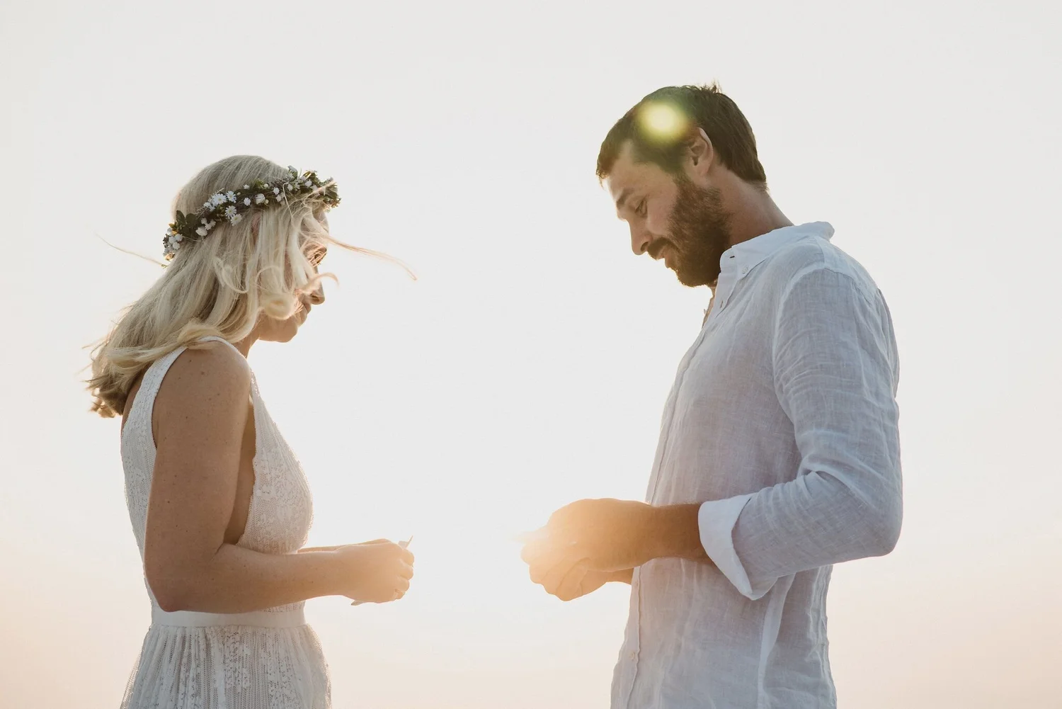 A couple is standing outdoors, facing each other, holding hands, during sunset. The woman has long blonde hair, a floral crown, and is wearing a white sleeveless dress. The man has dark hair, a beard, and is wearing a white button-up shirt.
