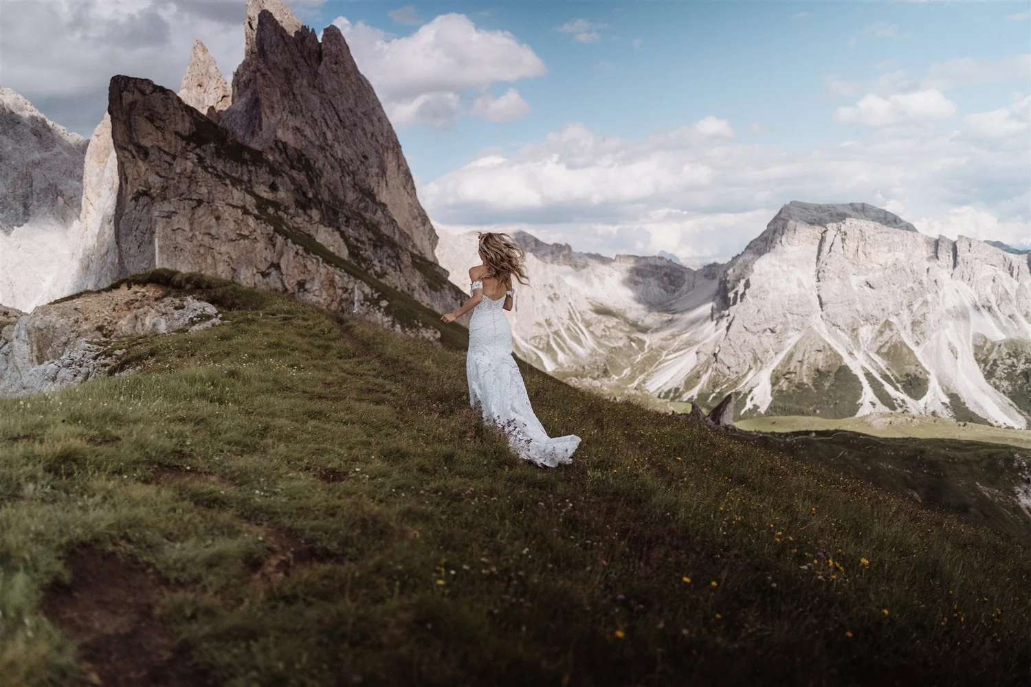 A woman in a wedding dress standing on a grassy hillside with mountains in the background.