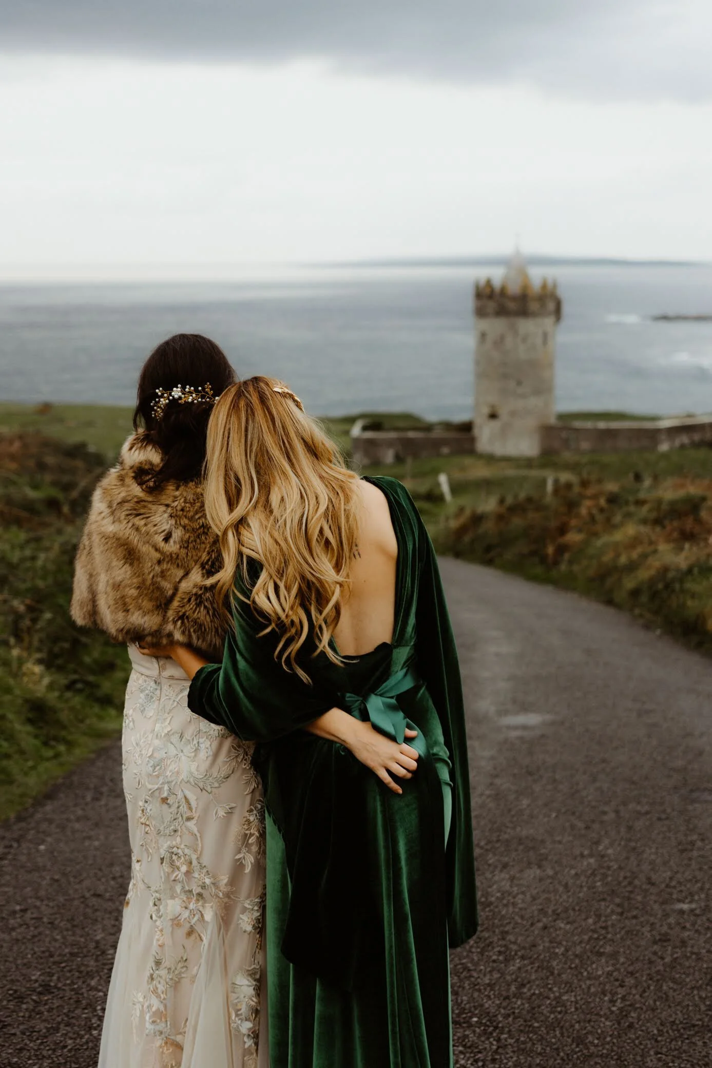 Two women stand together on a dark country road, overlooking a castle by the water under a cloudy sky. One woman wears a cream-colored dress with floral embroidery and lace, while the other wears a dark green velvet gown with an open back. The woman