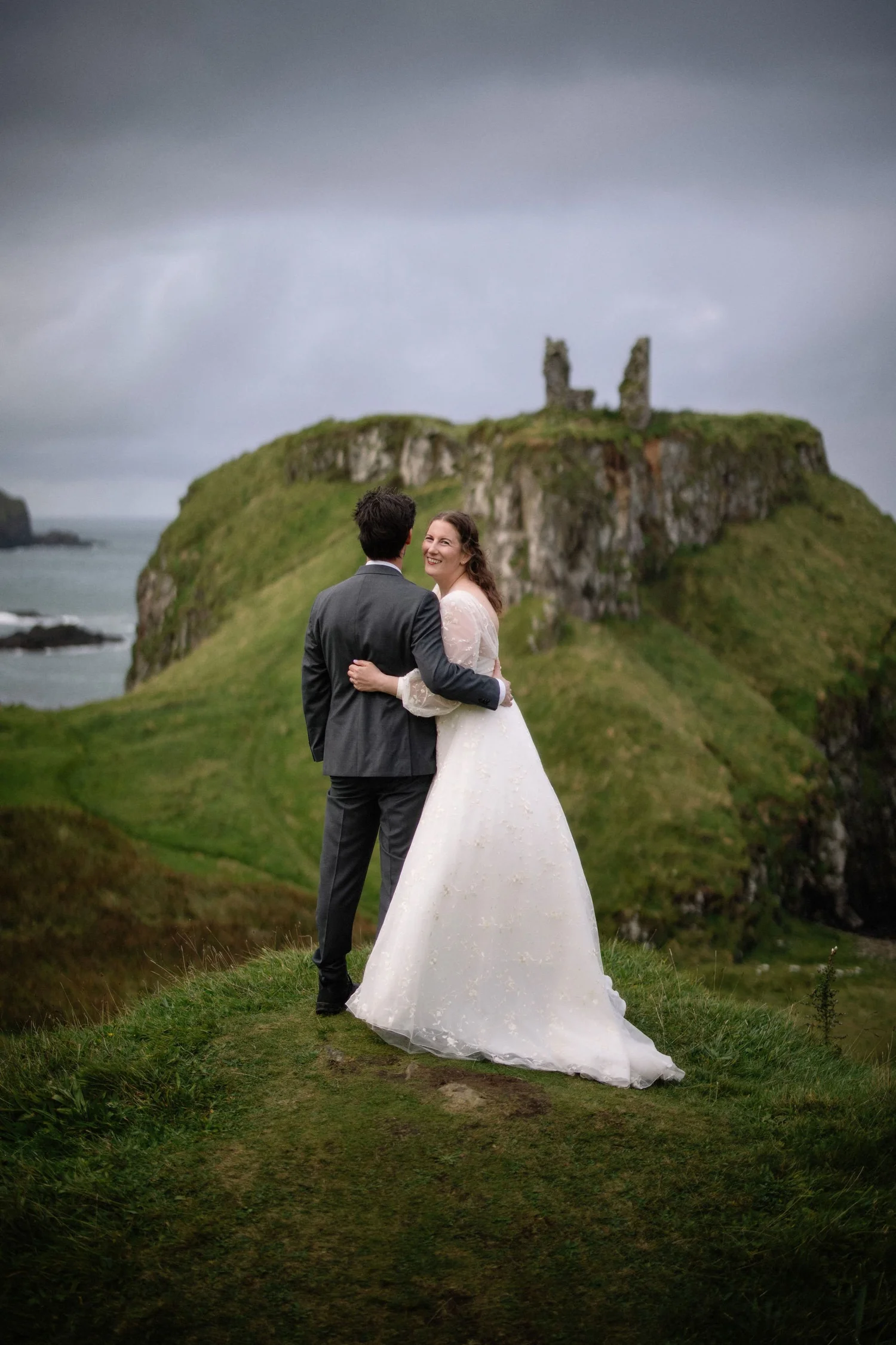 A bride and groom standing on a grassy path, holding each other and smiling. The bride is in a white wedding dress, and the groom is in a gray suit. Behind them is a green hillside with rocky formations and a medieval ruin on top, under a dark, cloud