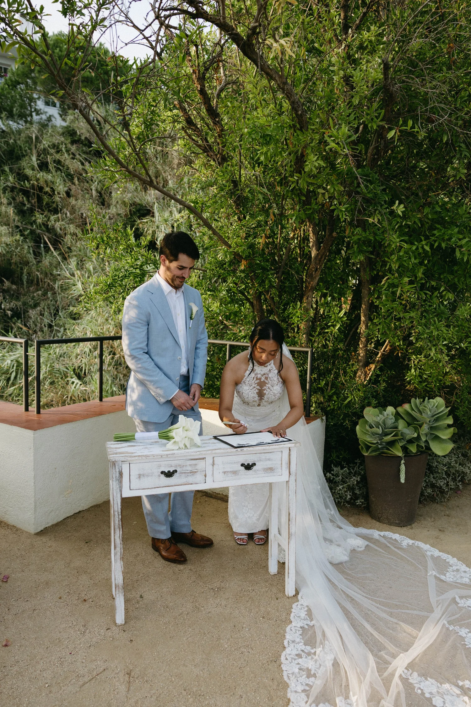 A couple signs a marriage certificate outdoors under a large tree with green foliage. The man wears a light blue suit and brown shoes, and the woman wears a white lace wedding dress with a long train. A white table with a bouquet of white calla lilie