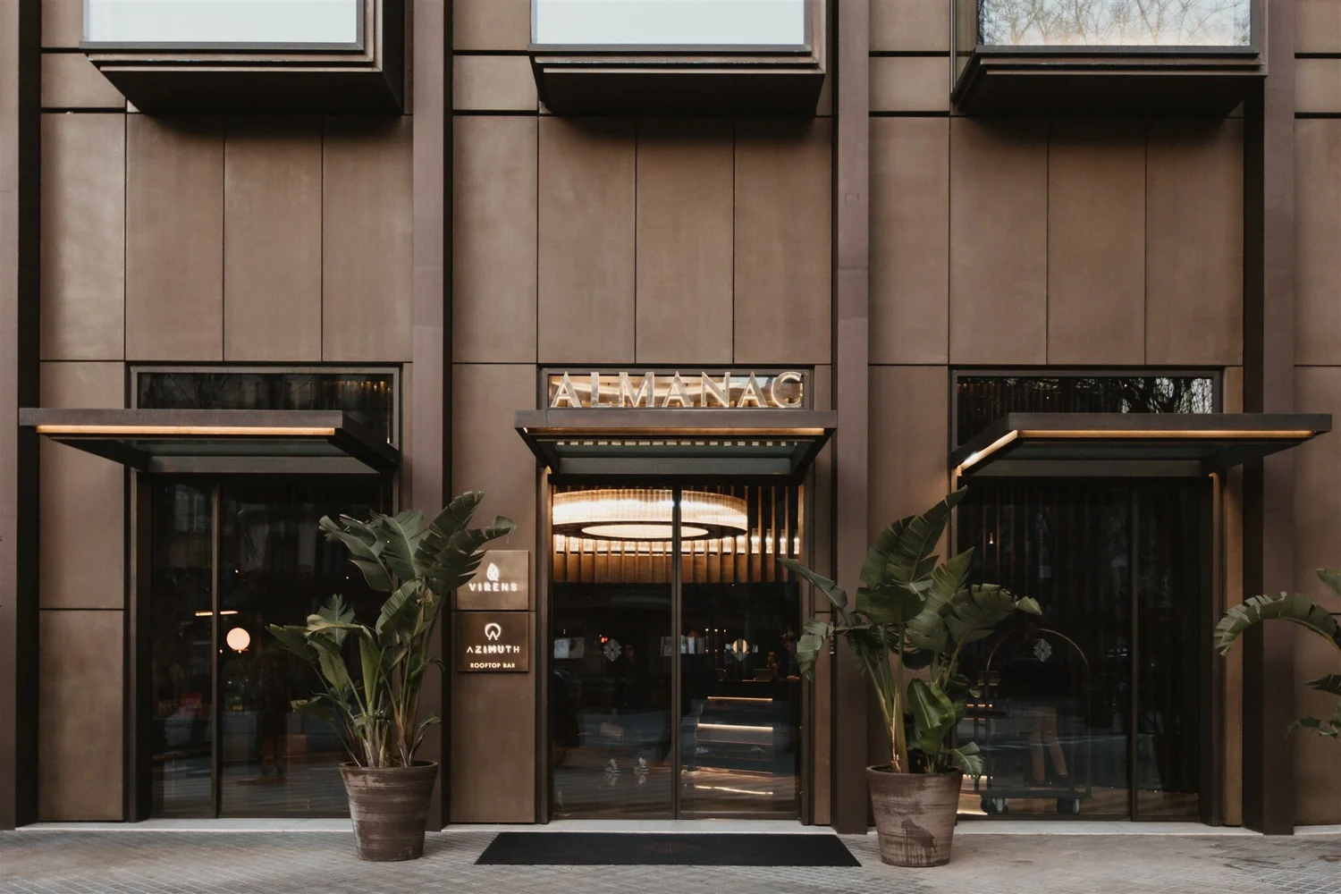 Storefront entrance of a building with large glass doors and windows, two potted plants flanking the entrance, and illuminated sign that reads 'ALMANAC'. The building has a modern design with framed panels on the facade.