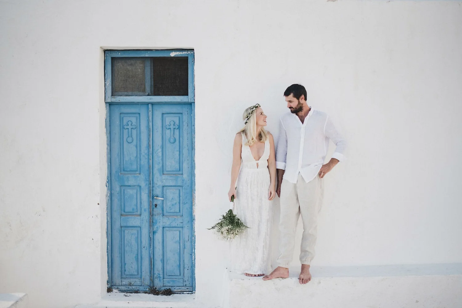 A man and woman dressed in white, smiling and standing barefoot against a white wall with a blue wooden door. The woman wears a floral headband and holds a bouquet, while the man has his hands in his pockets.