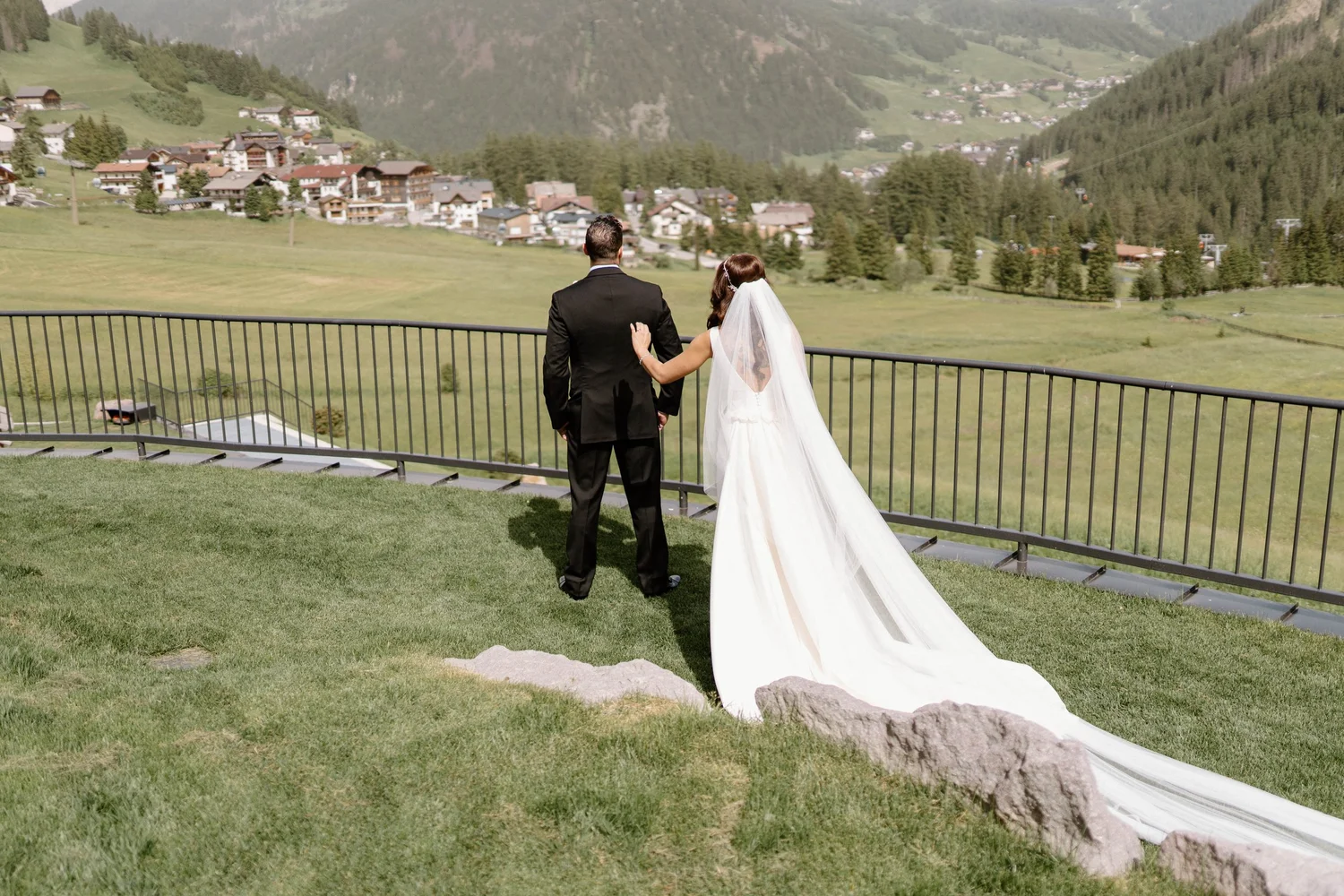 A bride and groom stand together on a grassy area overlooking a scenic mountain village, with the bride's veil flowing down and the groom dressed in a black suit.