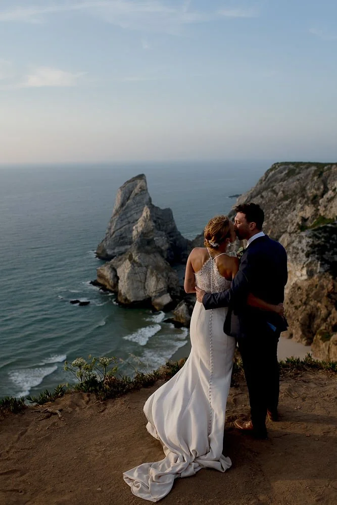 A bride and groom sharing a kiss on a cliffside overlooking the ocean, with rocky formations in the background.