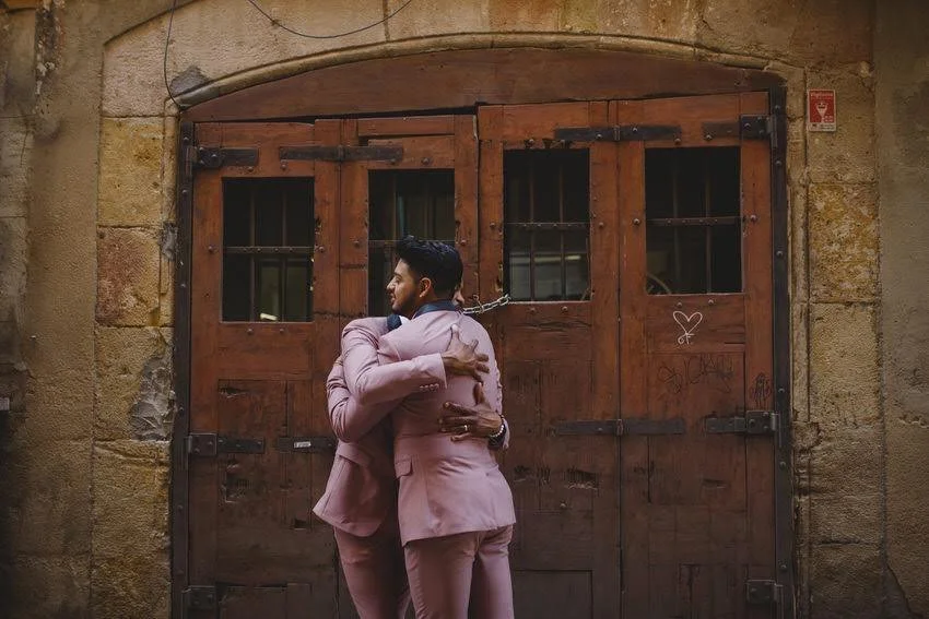Two men in pink suits hugging in front of a large wooden gate with metal hinges, set in a stone wall.