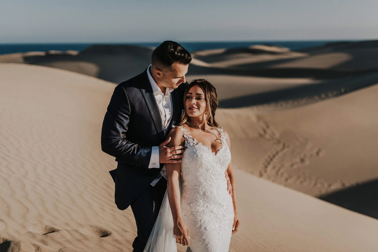 A bride and groom in wedding attire standing in a sandy desert landscape with dunes and a distant horizon.