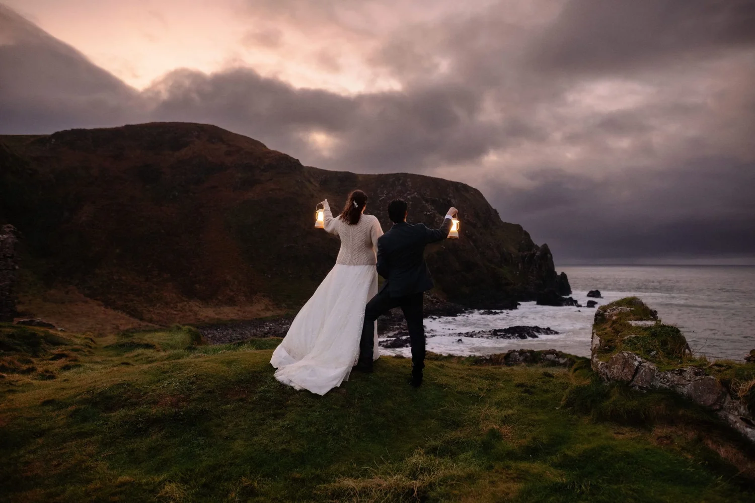 A couple, dressed formally, holding lanterns while standing on a grassy cliff overlooking the ocean at sunset or dusk, with cloudy sky overhead.