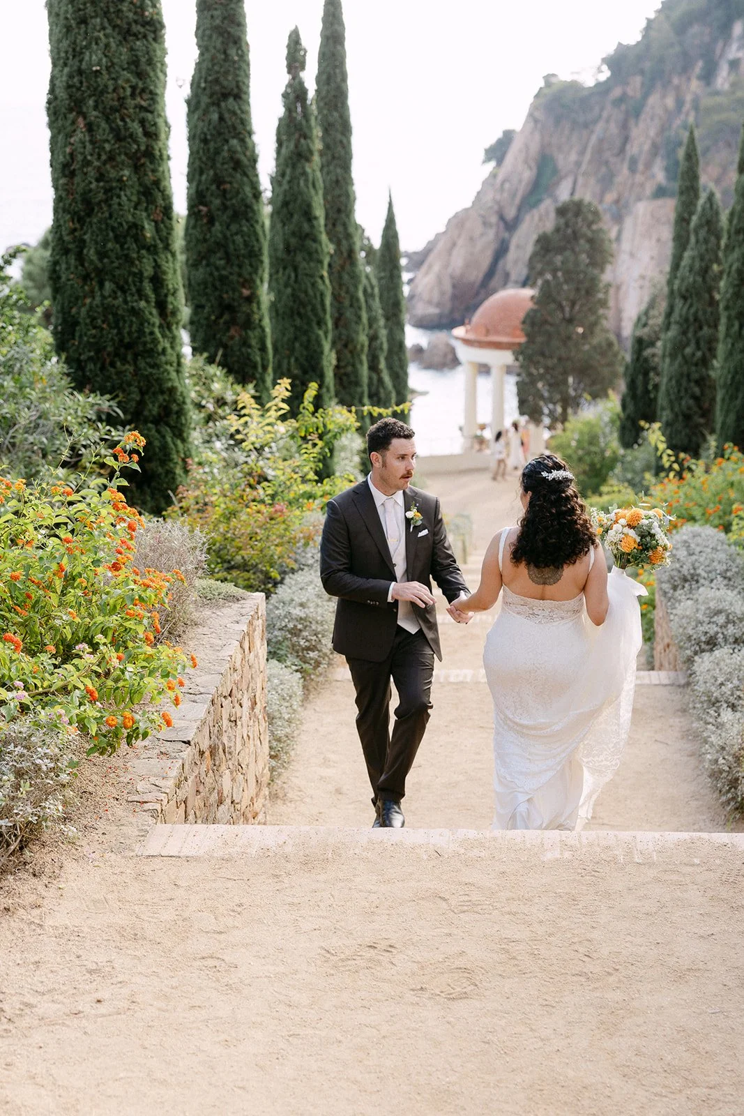 A groom and bride walking hand in hand along a garden path during their wedding, with tall cypress trees, colorful flowers, and a scenic backdrop of cliffs and the ocean.