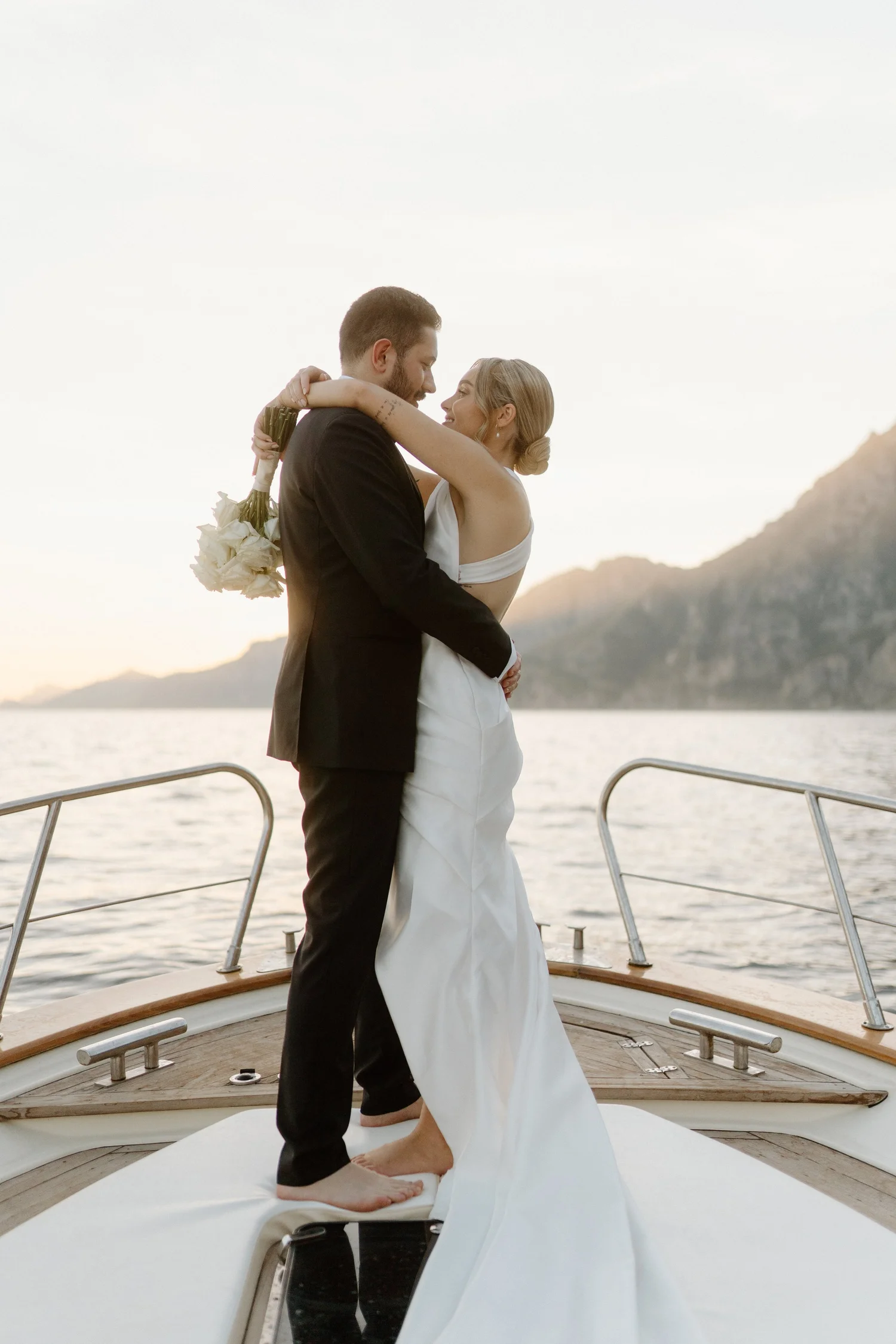 A newlywed couple embracing on a boat deck during sunset, with mountains and water in the background.