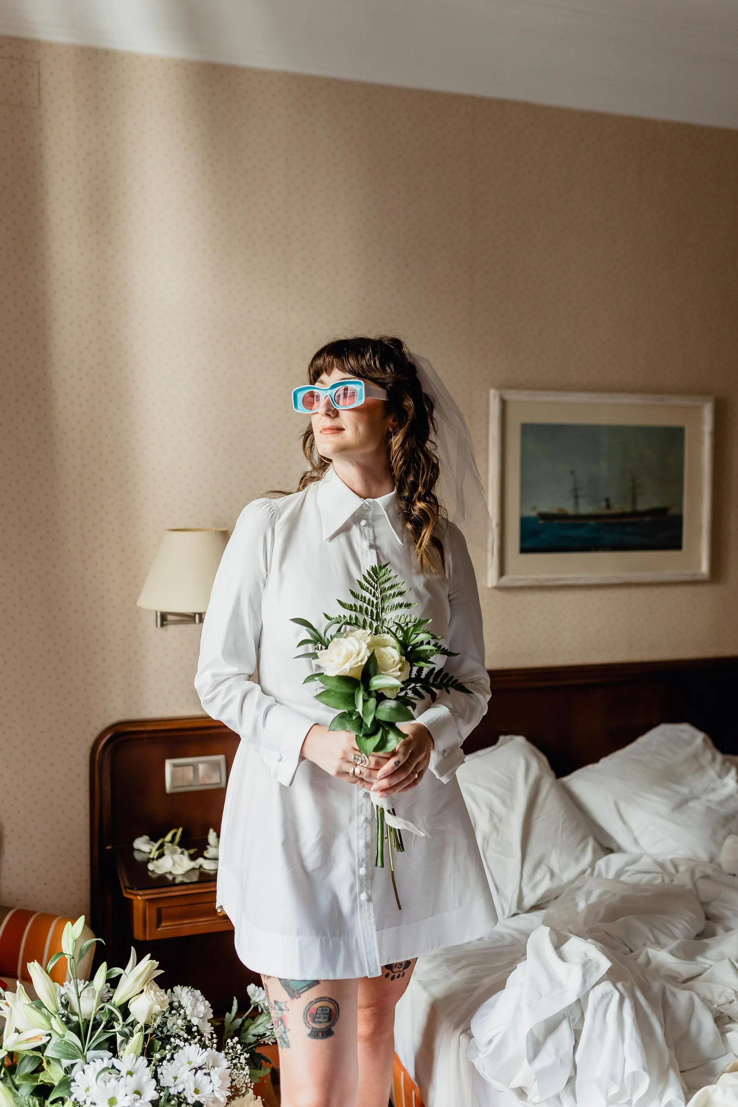 A woman dressed in a white shirt holding a bouquet of white roses and greenery, standing in a bedroom with unmade bed, floral wallpaper, and framed artwork of a ship on the wall.