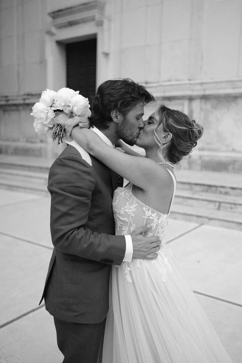 A black-and-white photo of a newlywed couple sharing a kiss, with the bride holding a bouquet of flowers, indoors with classical architecture.