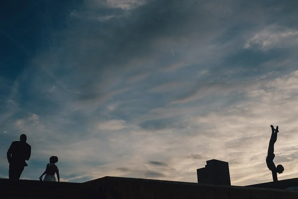 Silhouettes of three people standing on a rooftop with a cloudy sky in the background, including a person performing a handstand.